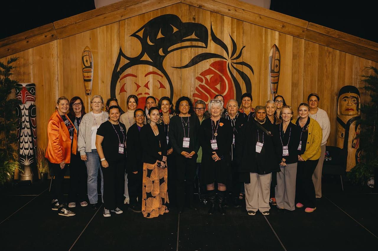 The Australian delegation of Indigenous women at the IWI Indigenous Women in Industry Summit in Vancouver, standing together on stage in front of a carved wooden longhouse front with a Thunderbird des